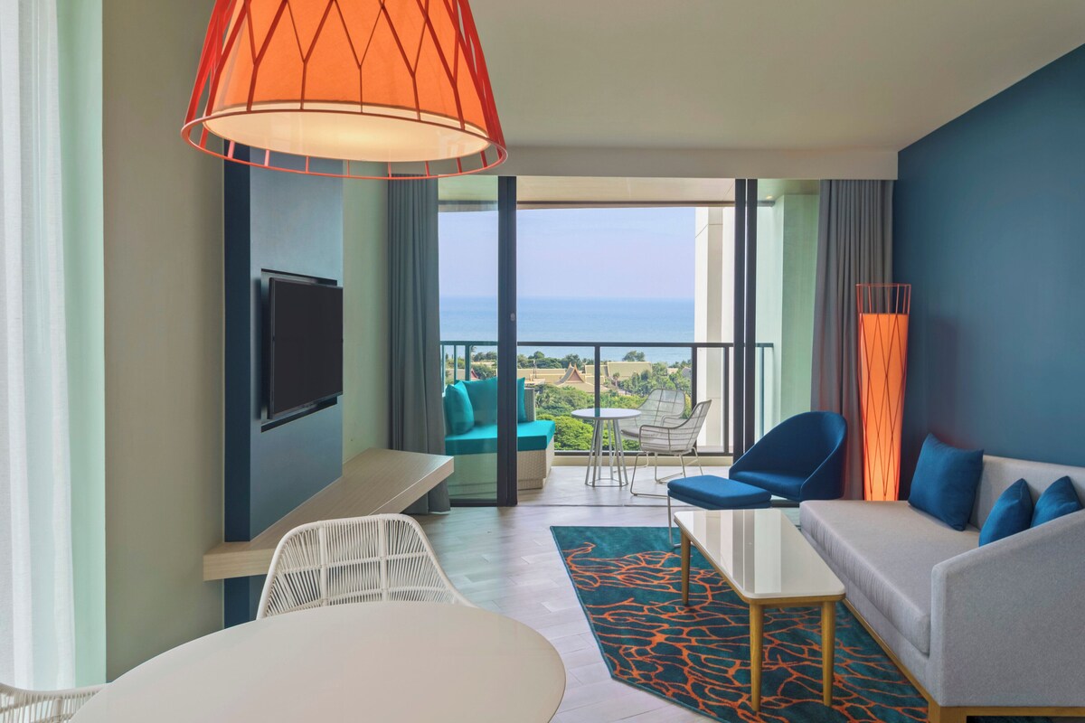 Quarto de hotel moderno com varanda, televisão de tela plana, sofá, mesa de jantar e tapete estampado.