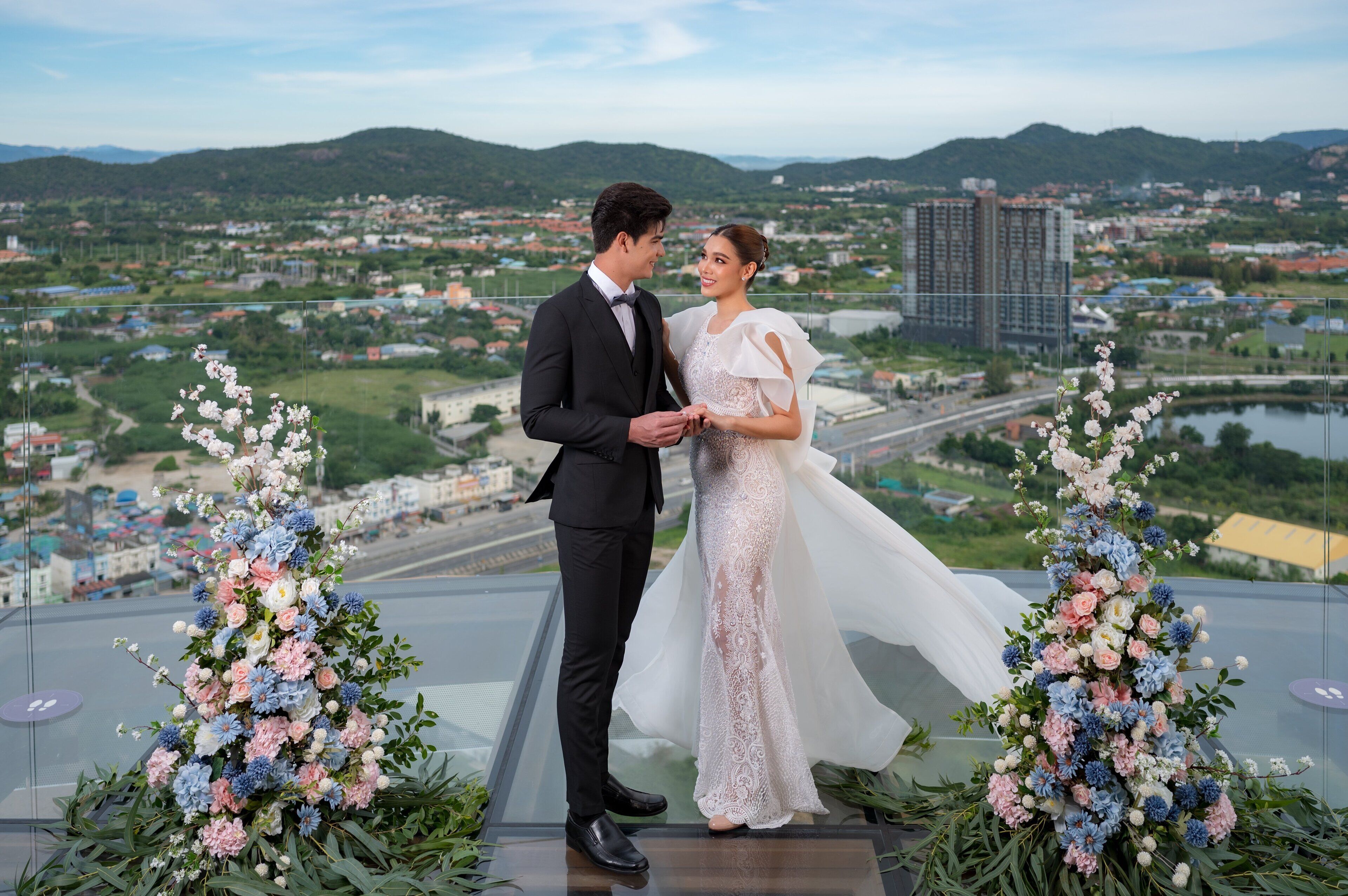 Un couple se tient sur un balcon, avec une vue sur la ville et des montagnes en arrière-plan.