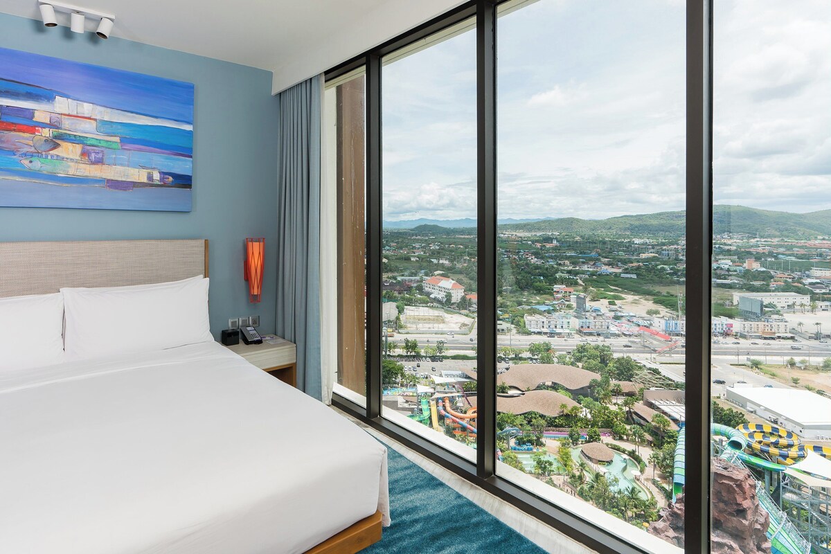 Quarto de hotel com uma cama grande, um quadro na parede, vista para a cidade através de uma janela ampla e uma mesa de cabeceira com telefone e abajur.