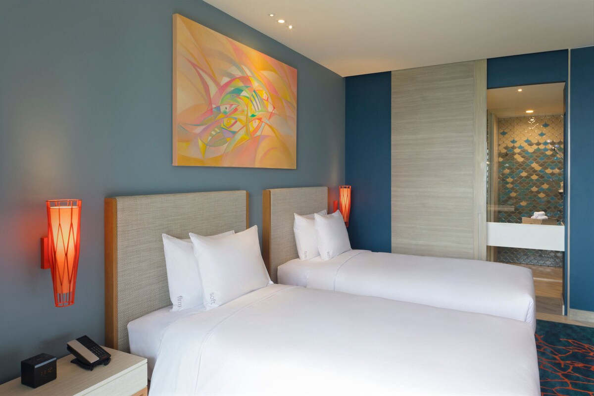 Quarto de hotel com duas camas, uma pintura abstrata grande, uma mesa de cabeceira com telefone e um banheiro com chuveiro azulejado.