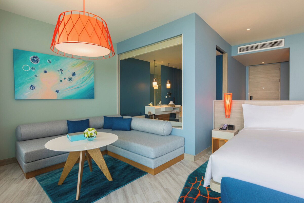 Quarto de hotel moderno com sofá, cama, mesa redonda e banheiro visível por uma porta aberta.