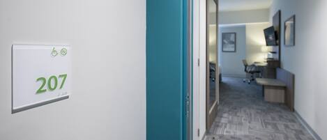 A hallway with a sign indicating room number 207, a teal-colored door, and a glimpse of a workspace area with a desk and chair.
