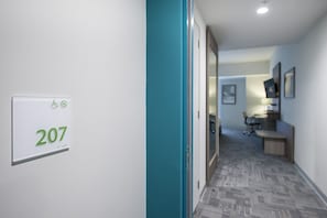 A hallway with a sign indicating room number 207, a teal-colored door, and a glimpse of a workspace area with a desk and chair.