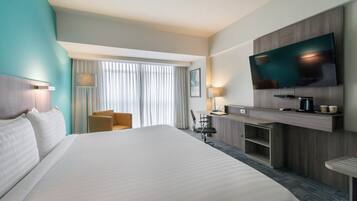 A hotel room with a large bed, a desk with a flat-screen TV, a chair, and a desk lamp.