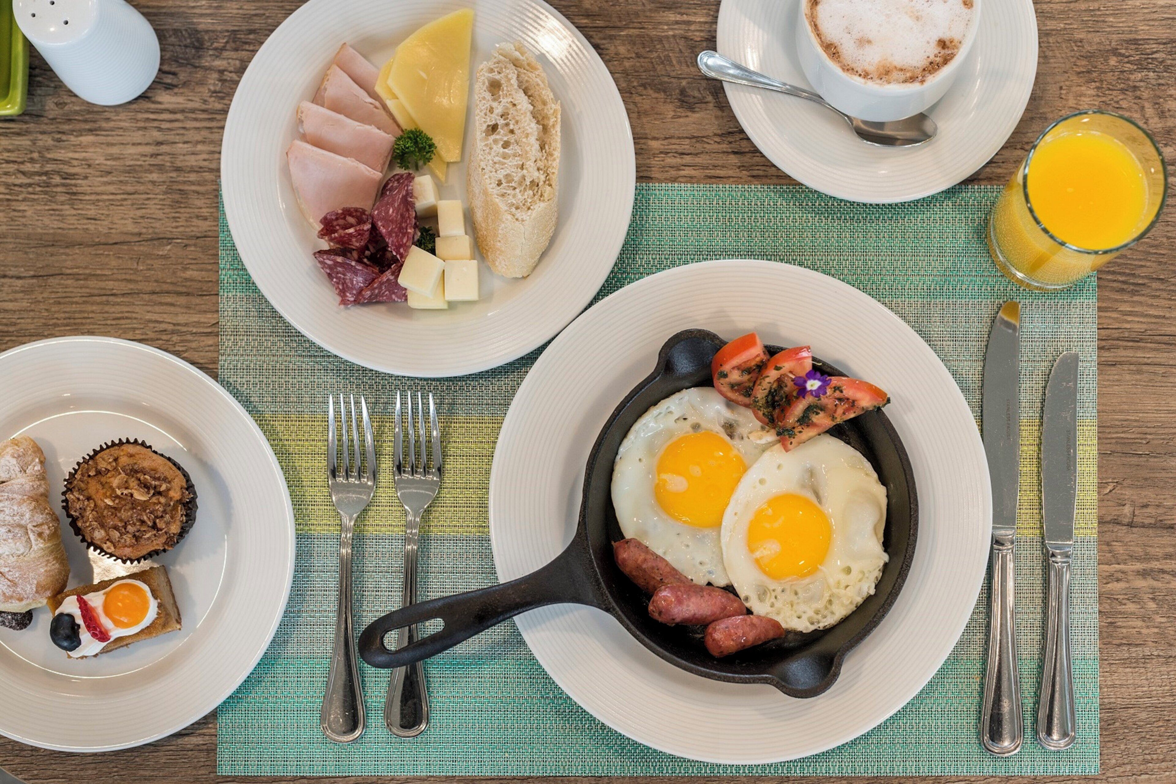 A breakfast setting with plates of eggs, sausages, and toast, a skillet with eggs, and a glass of orange juice.