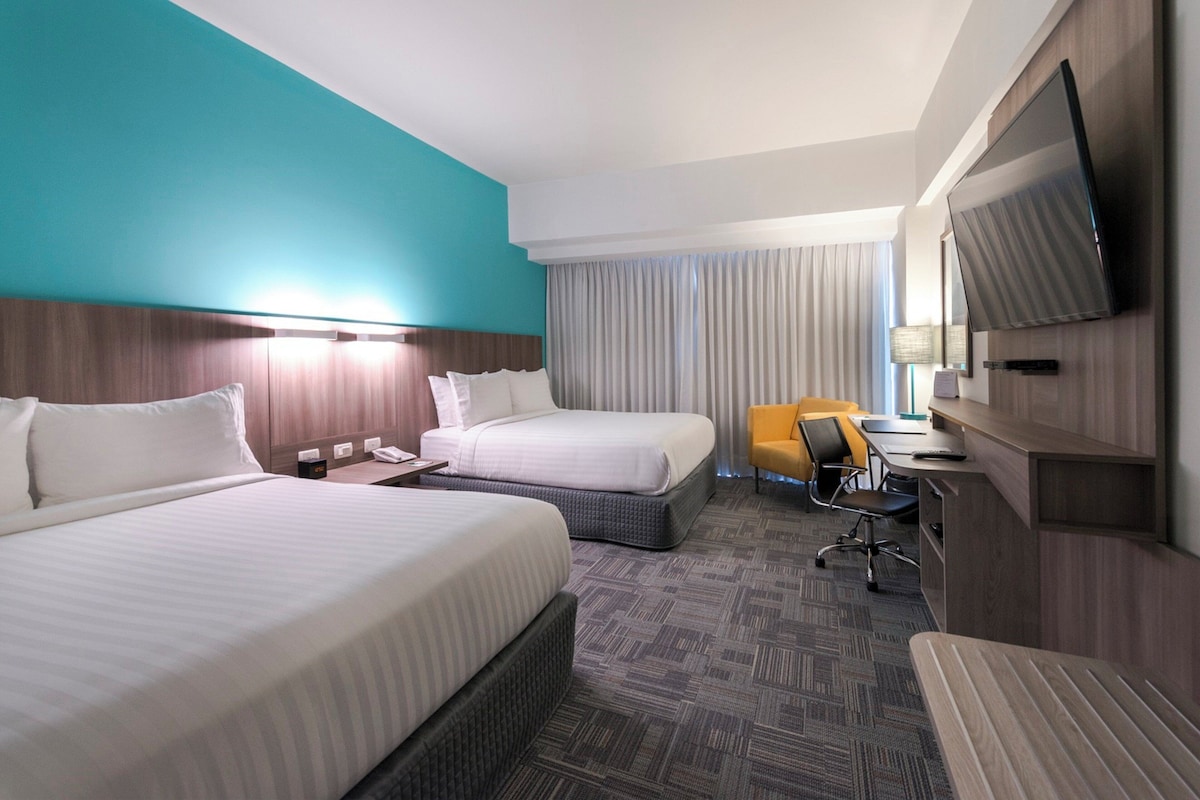 A hotel room with two beds, a desk, a chair, a television, and a large window with curtains.