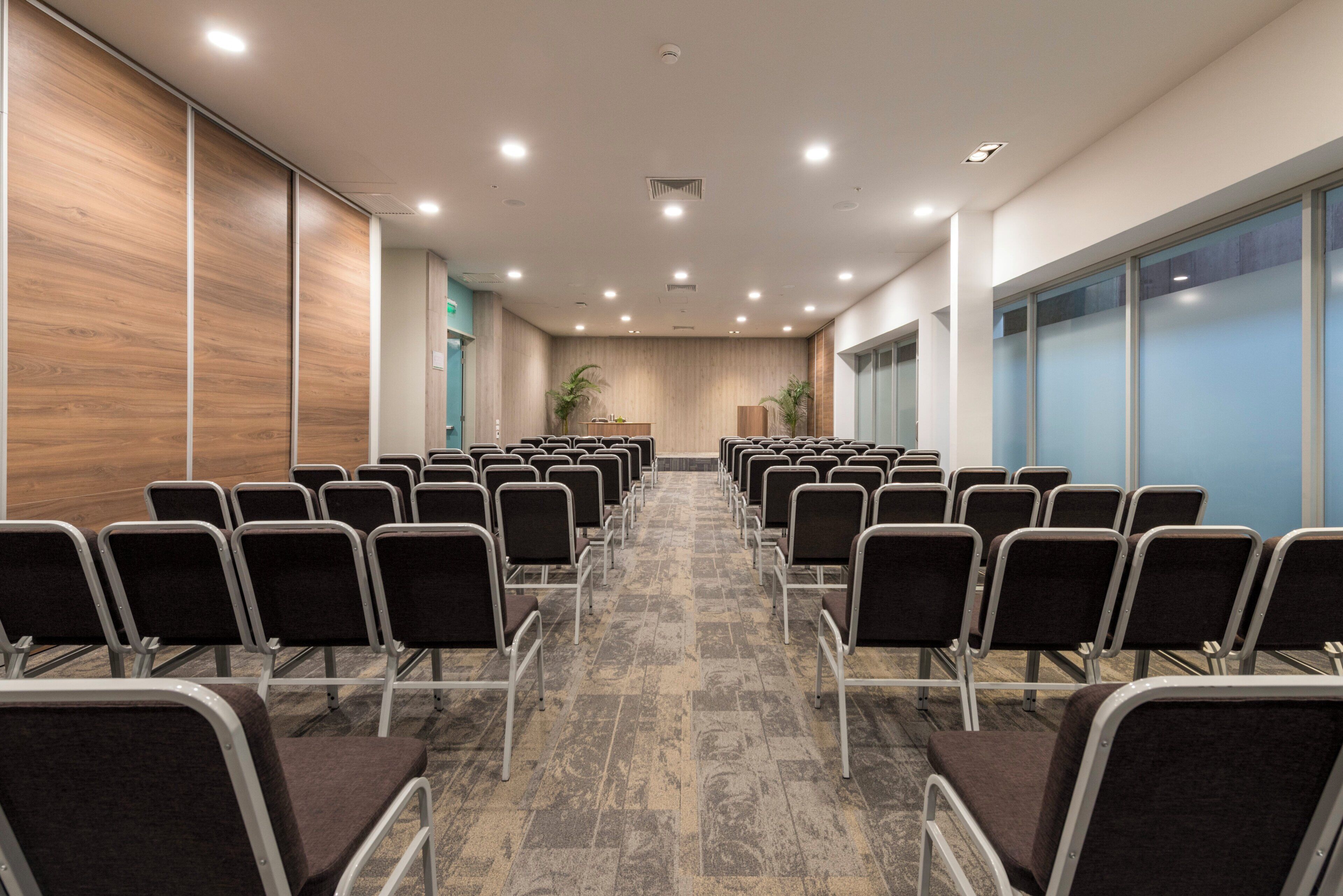 Ein Konferenzraum mit Reihen von Stühlen, einer Holzvertäfelung, einer blauen Wand mit Fenstern und einem gemusterten Boden.