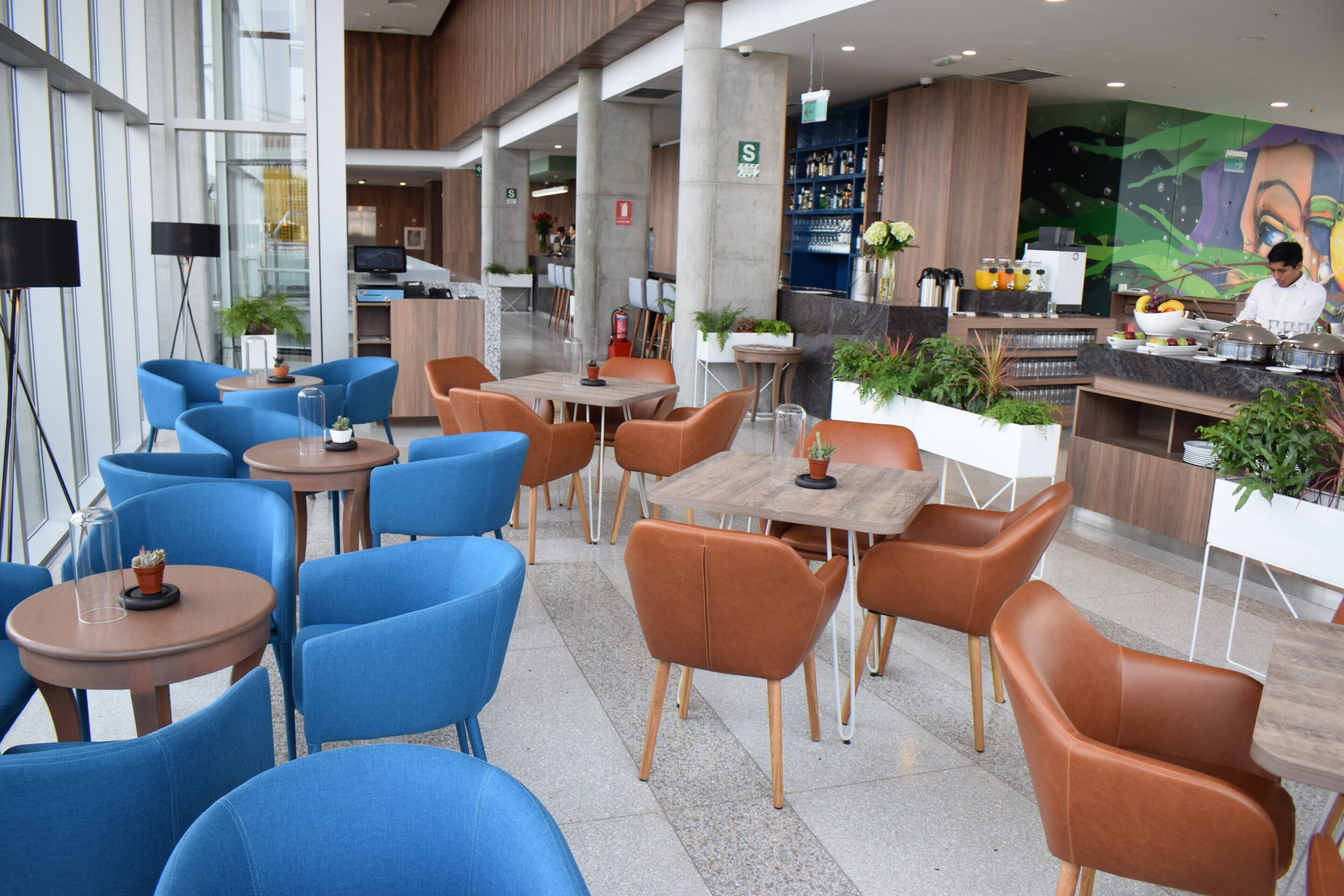 A modern indoor seating area with blue and brown chairs, tables, and a bar area with a chef preparing food.