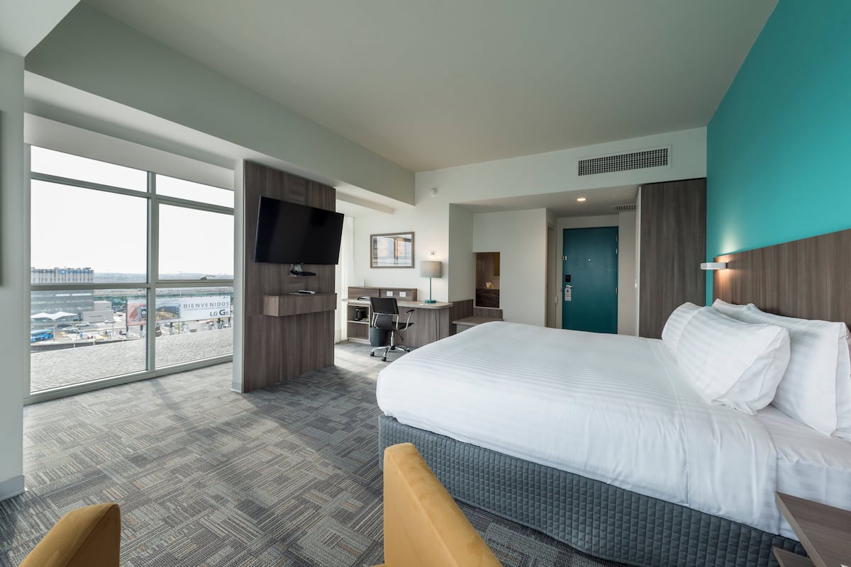 A hotel room with a large bed, a desk, a chair, a TV, and a view of the city.