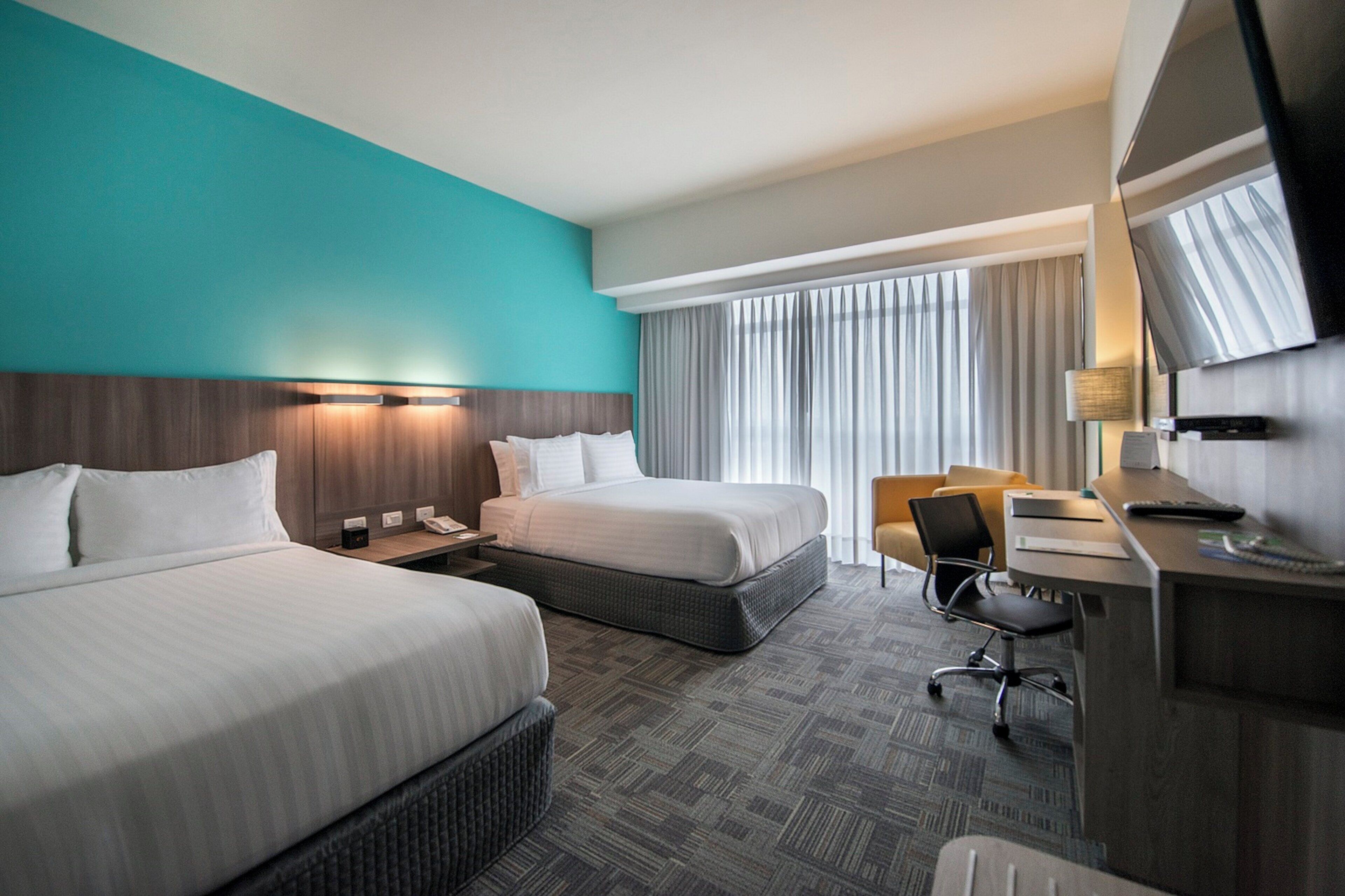 A hotel room with two beds, a desk, a chair, a TV, and a window with curtains.