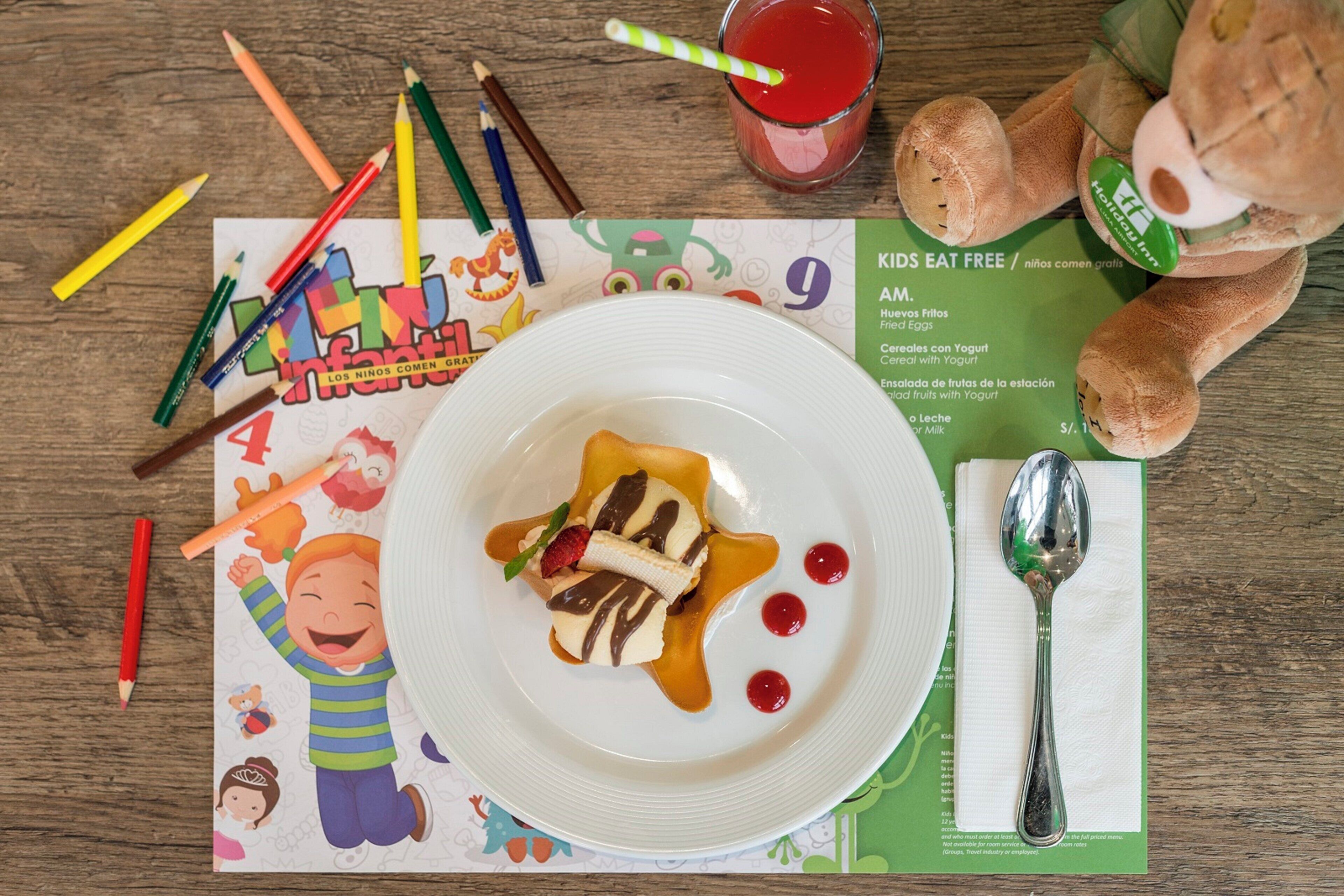 Refeição infantil com prato de comida, uma colher, um copo de bebida vermelha e um cardápio sobre uma mesa de madeira.