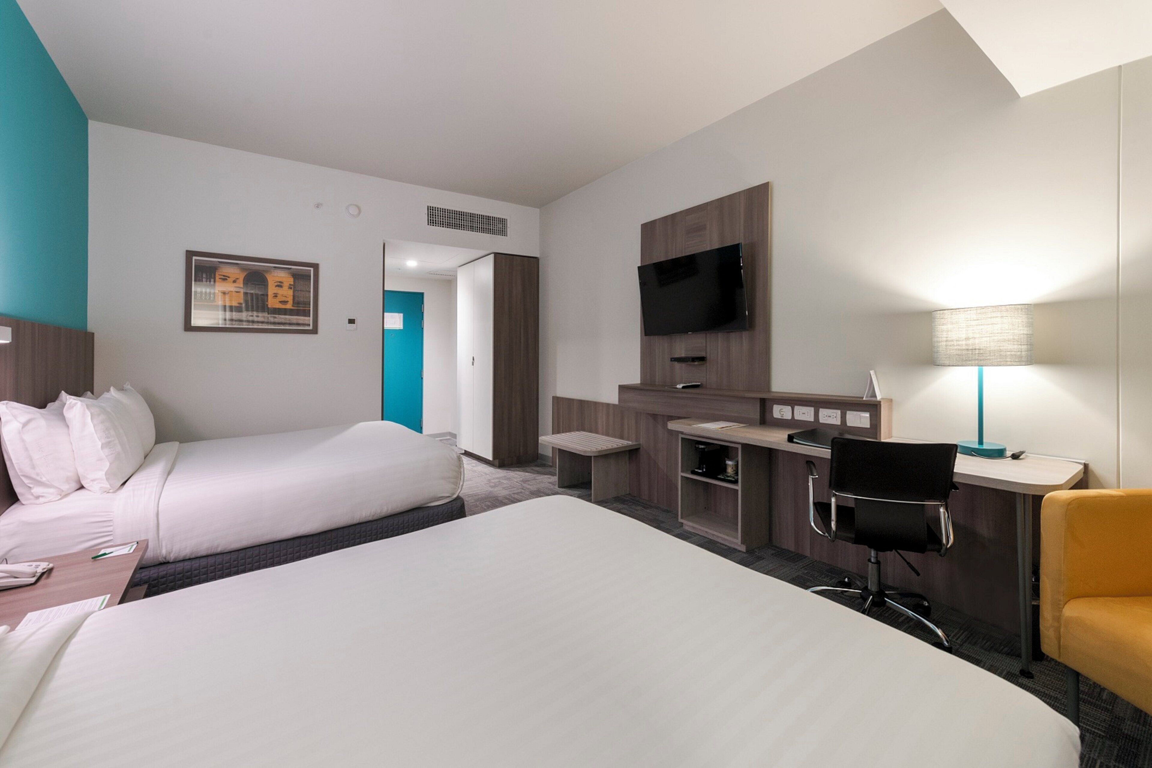 A hotel room with two beds, a desk, a chair, a TV, and a lamp.