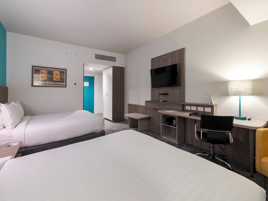 A hotel room with two beds, a desk, a chair, a TV, and a lamp.