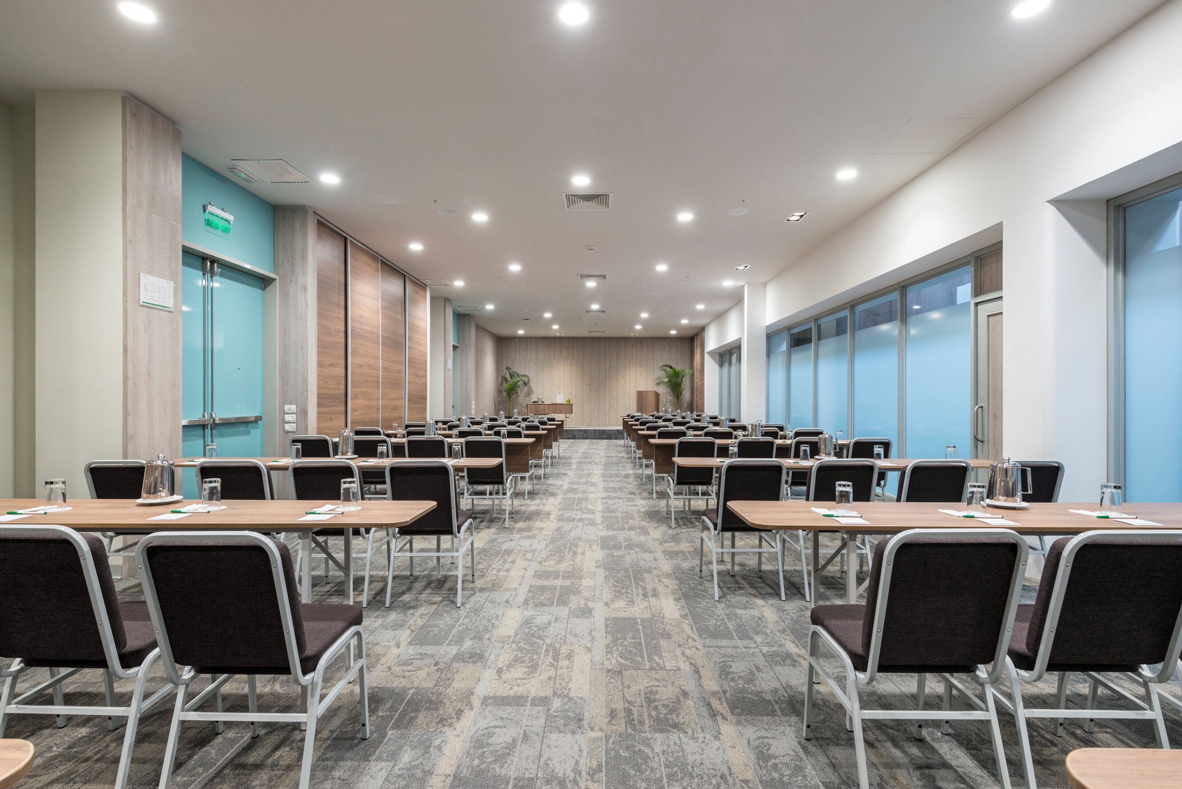 A conference room with rows of tables and chairs, a wooden accent wall, and a large window on the right.