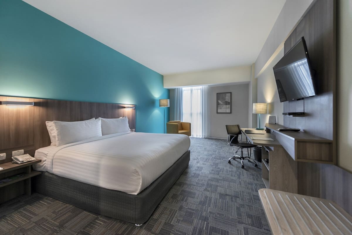 A hotel room with a large bed, a desk, a chair, a TV mounted on the wall, and a window with curtains.