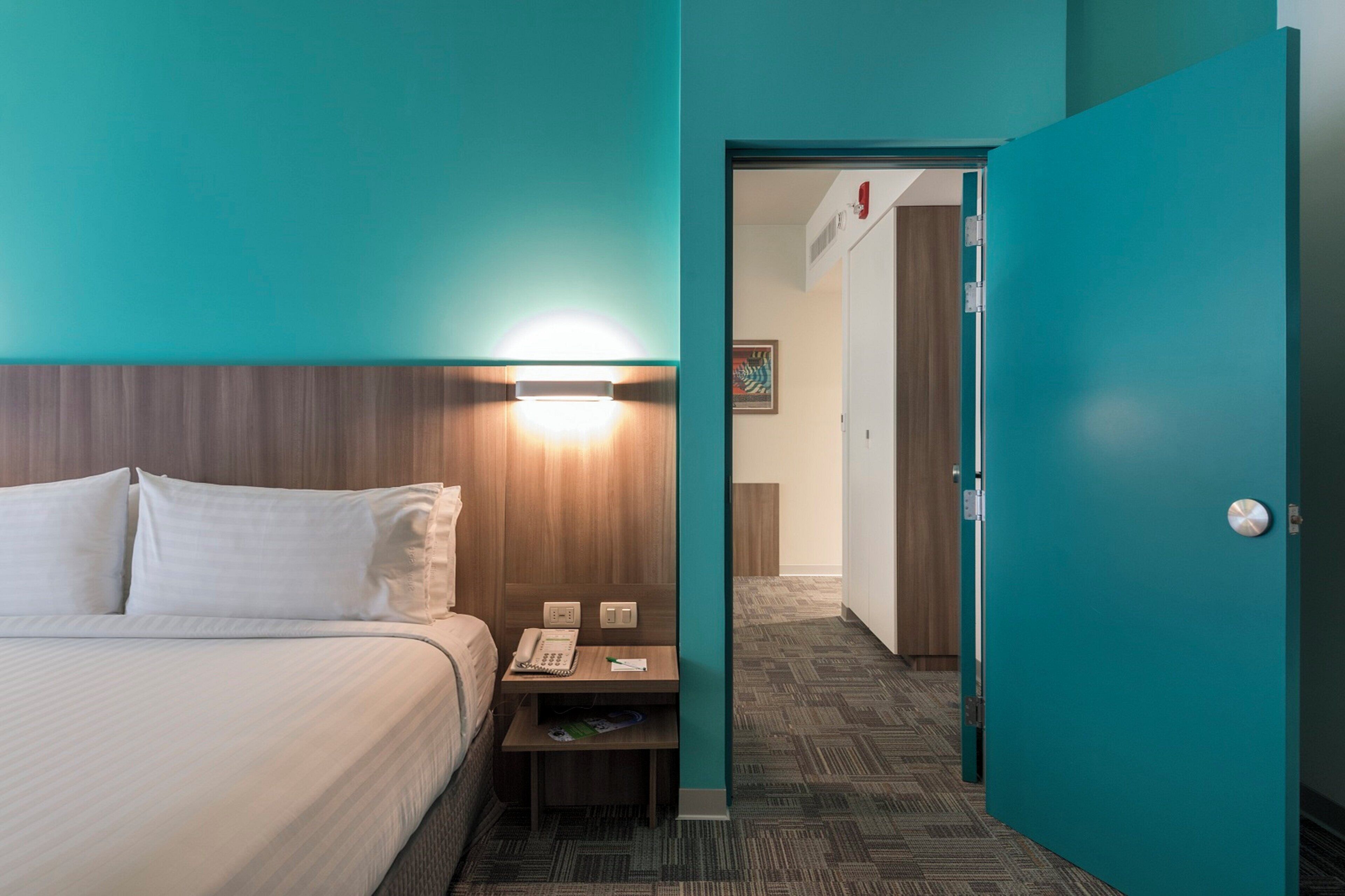 A hotel room with a bed, bedside table, lamp, and a door leading to another room.