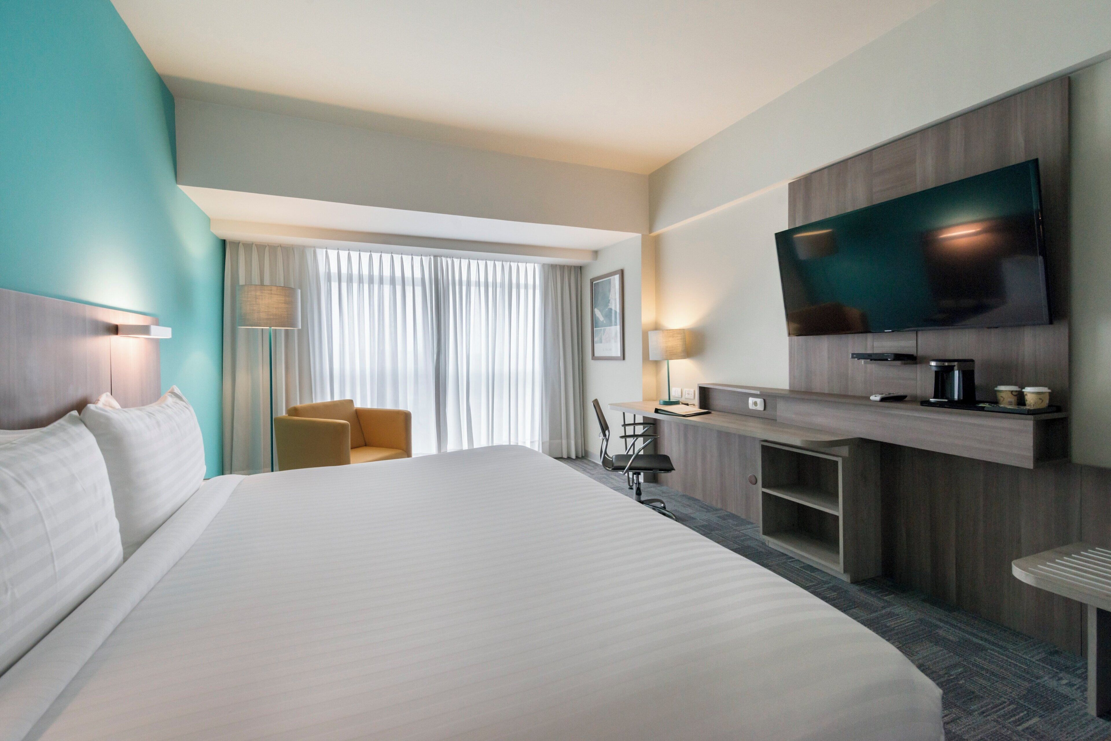 A hotel room with a large bed, a desk with a flat-screen TV, a chair, and a desk lamp.