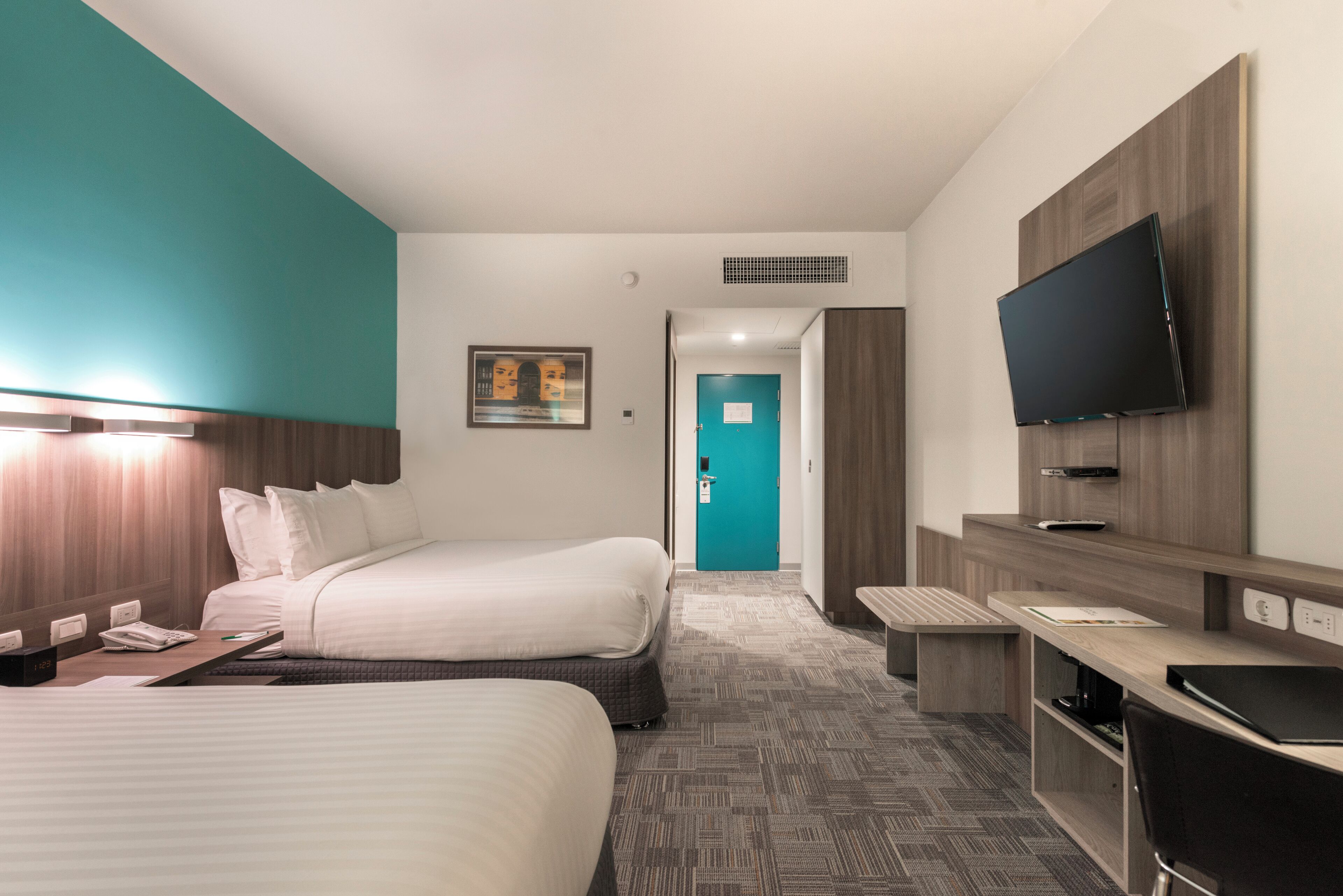 A hotel room with two beds, a desk, a flat-screen TV mounted on the wall, and a bathroom door in the background.
