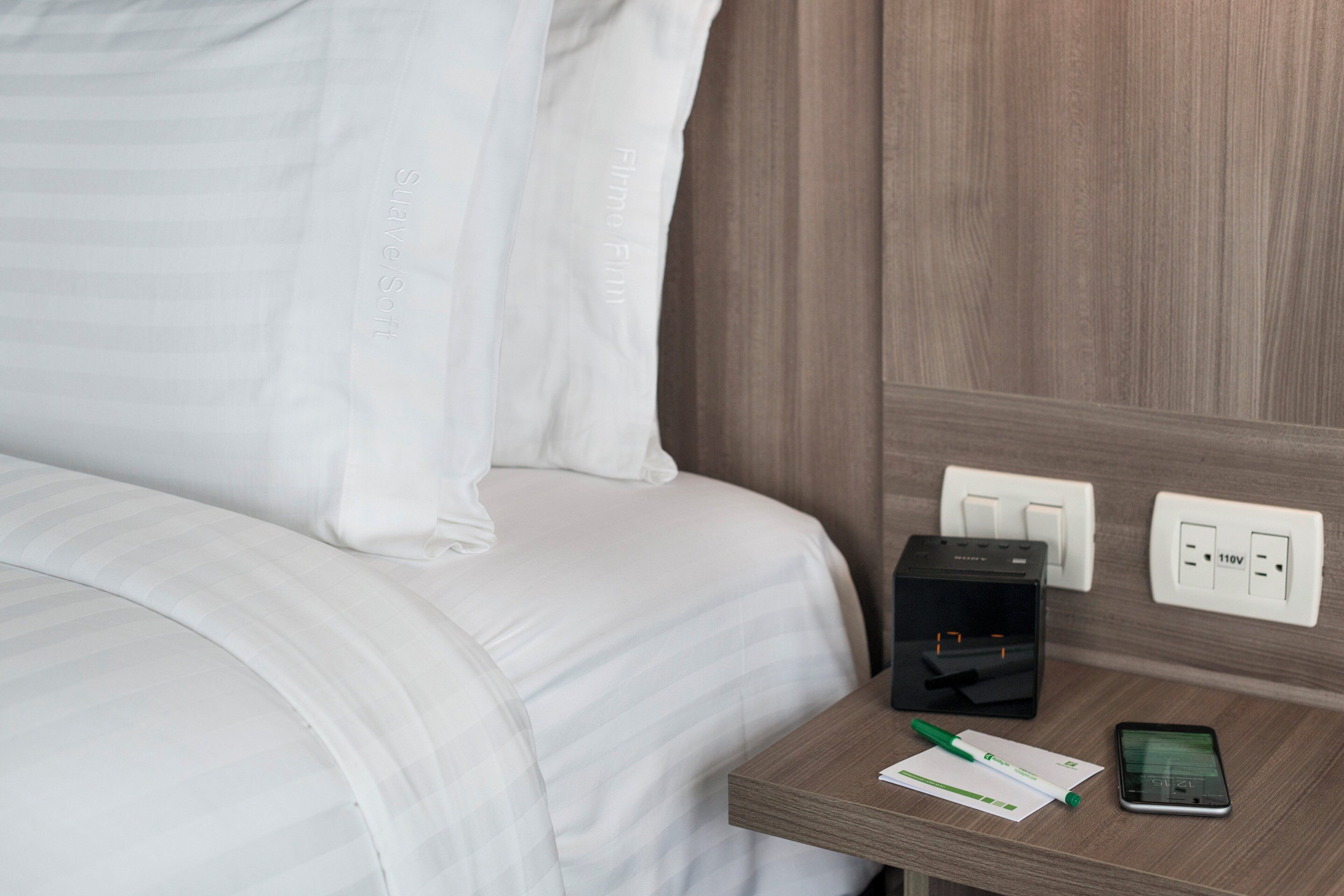 Una cama bien hecha con ropa blanca, una mesita de noche con un adaptador de corriente, un teléfono inteligente y una pluma.