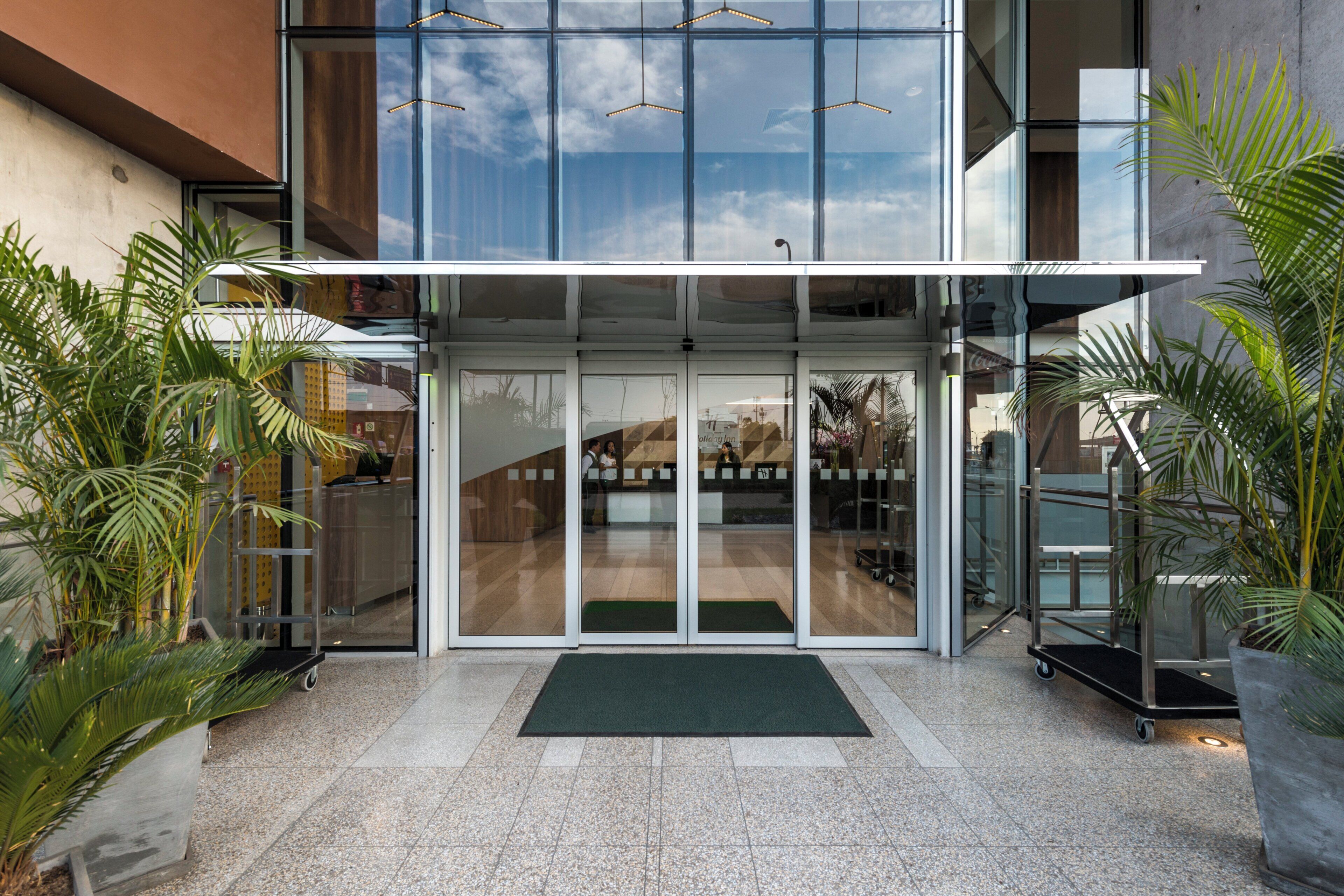 Entrada de hotel con puerta de vidrio, felpudo verde y plantas en macetas.