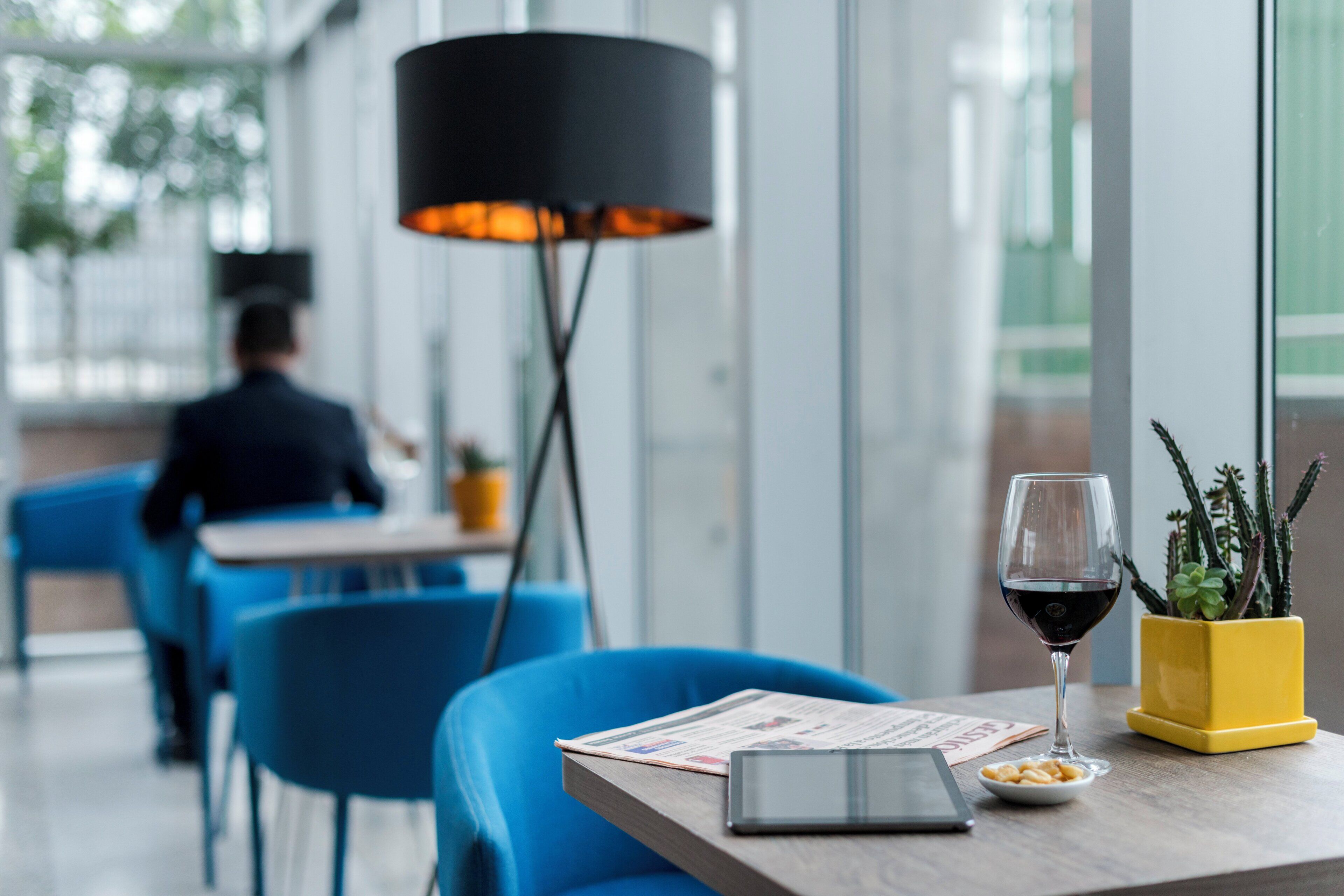 Interior de un restaurante con sillas azules, una mesa con una copa de vino, una tableta y una planta amarilla.