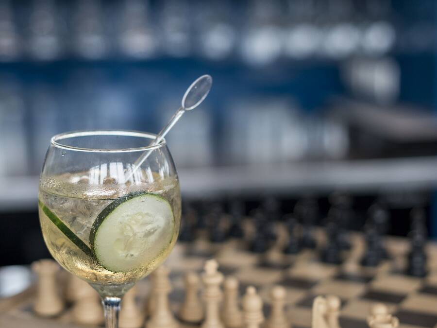 Un vaso de una bebida transparente con una rodaja de pepino y una pajita, sobre una superficie de mármol con un tablero de ajedrez al fondo.