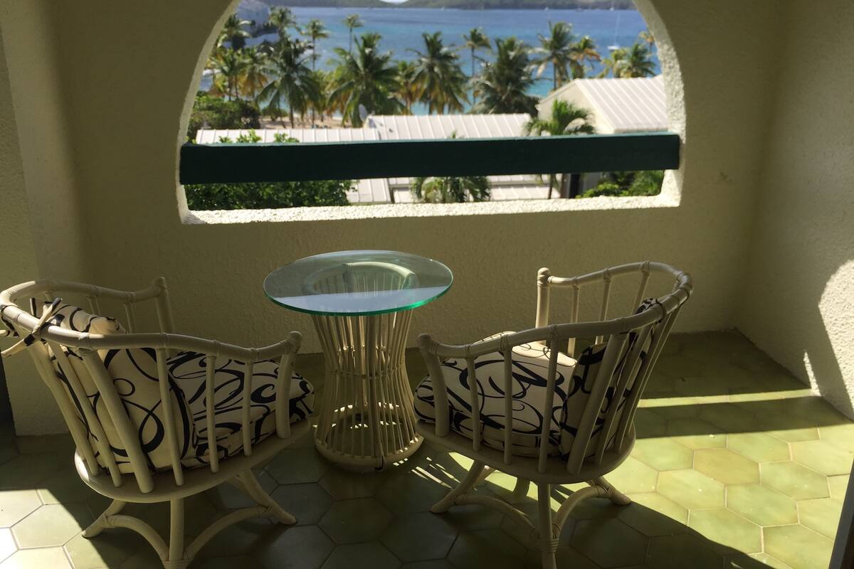 Uma varanda com duas cadeiras e uma mesa, com vista para uma praia com palmeiras e o mar.
