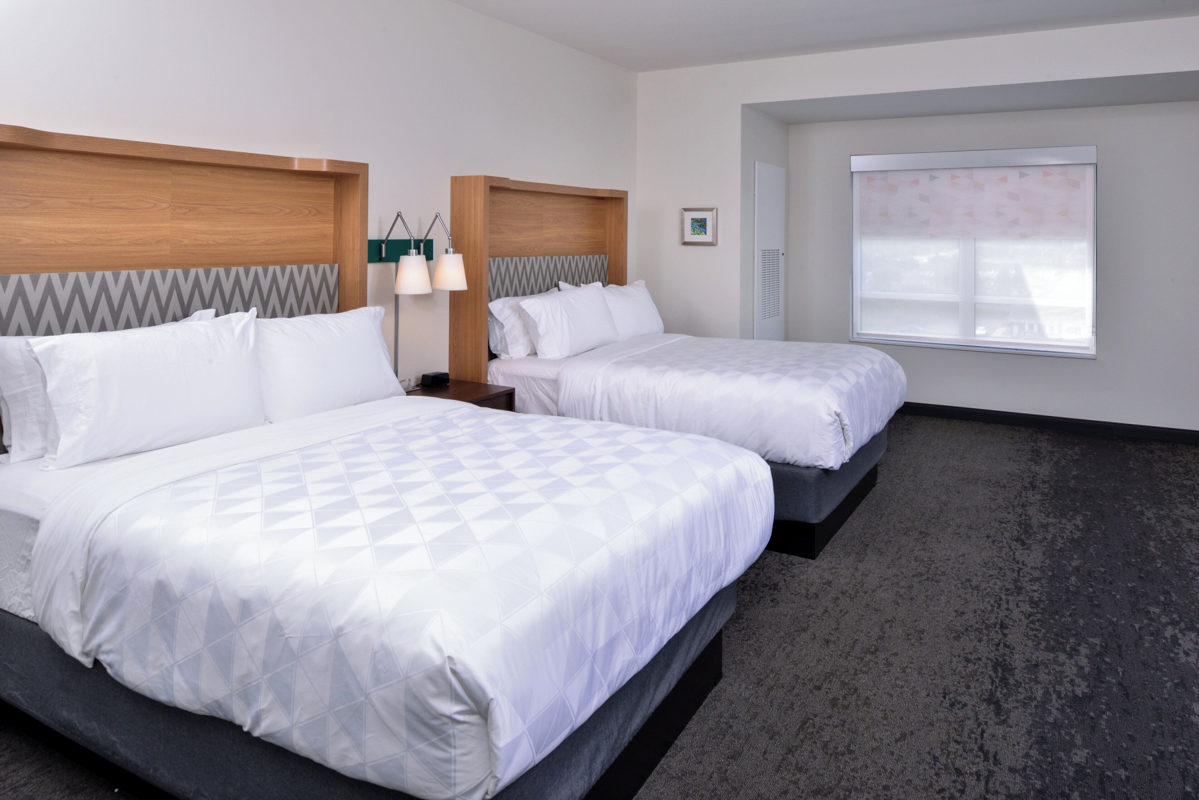 A hotel room with two beds, a nightstand, a lamp, a window with blinds, and a picture on the wall.