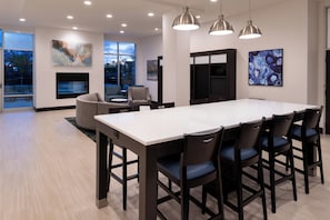 A modern kitchen with a large island, dark chairs, pendant lights, and artwork on the walls.