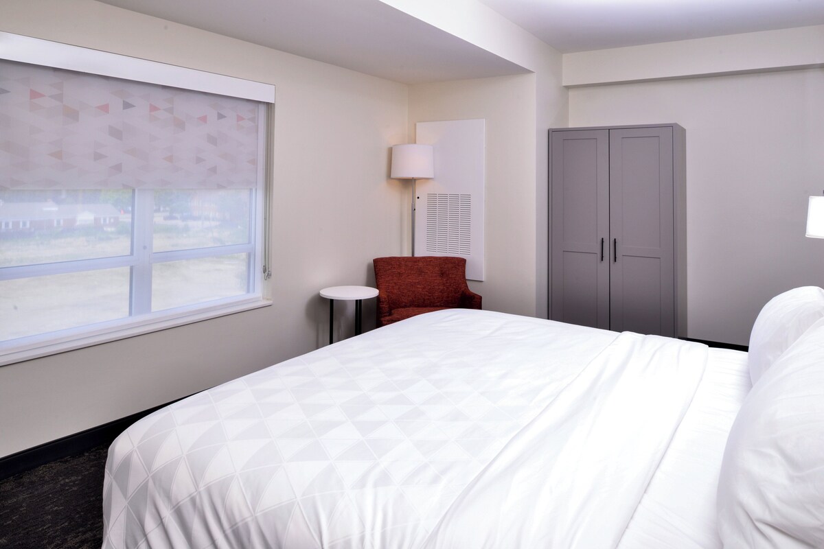 A hotel room with a large bed, a red chair, a wardrobe, and a window with blinds.