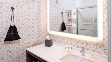 Un baño con papel tapiz estampado, un espejo grande, un lavabo con artículos de aseo y una bolsa colgada.