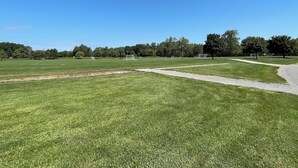 A large, open grassy field with multiple soccer goals and a paved path.