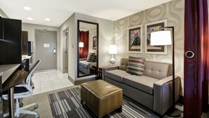A hotel room with a desk, chair, sofa, ottoman, and a TV. - Home2 Suites by Hilton KCI Airport (Kansas City)