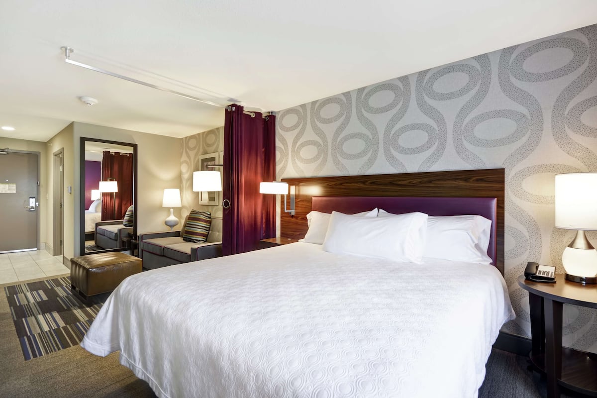 Une chambre d’hôtel avec un grand lit, deux tables de chevet avec des lampes, une tête de lit en bois, un miroir, un coin salon avec une chaise et une petite table, et un mur décoré d’un motif.