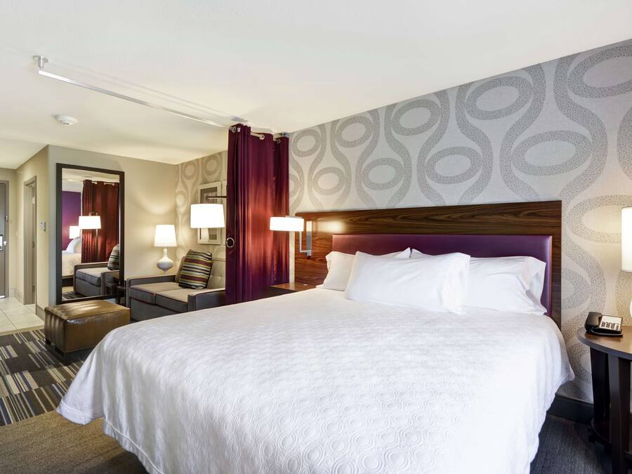 A hotel room with a large bed, two bedside tables with lamps, a wooden headboard, a mirror, a seating area with a chair and a small table, and a patterned wall.