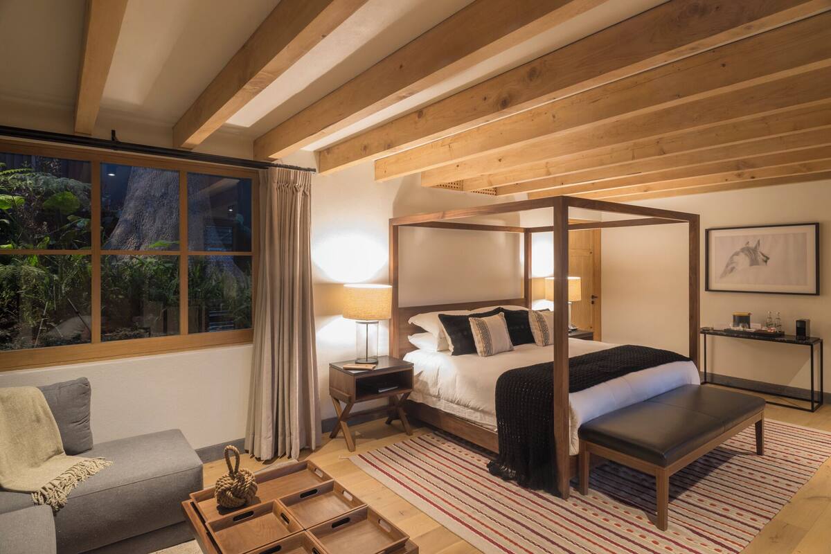 Ein Schlafzimmer mit einem Himmelbett, einem Nachttisch, einer Lampe, einer Bank, einem Sofa und einem Fenster mit Vorhängen.