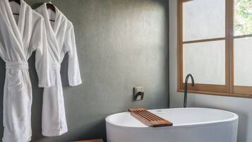 A modern bathroom with a freestanding bathtub, wooden bench, and two white robes hanging on the wall.
