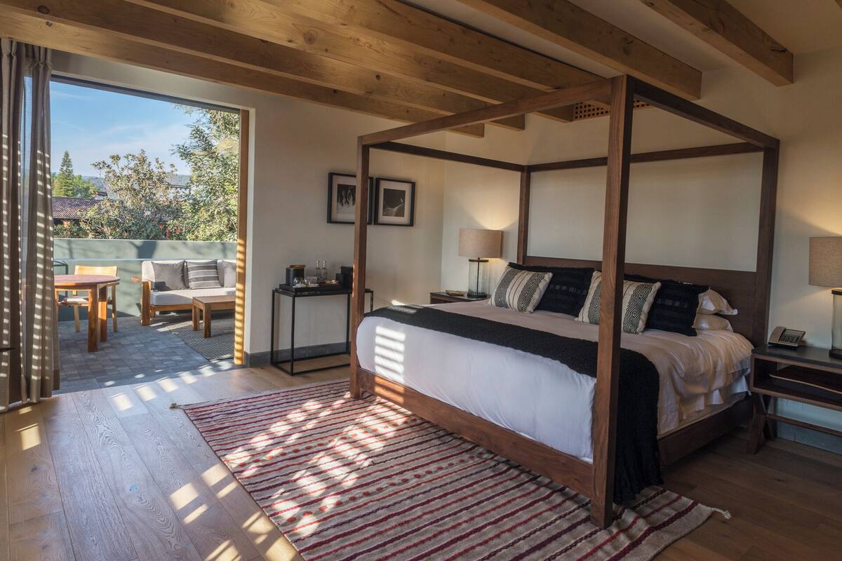 Ein Schlafzimmer mit Himmelbett, Holzboden, einem Streifenmuster-Teppich und Blick ins Freie durch eine Schiebetür.