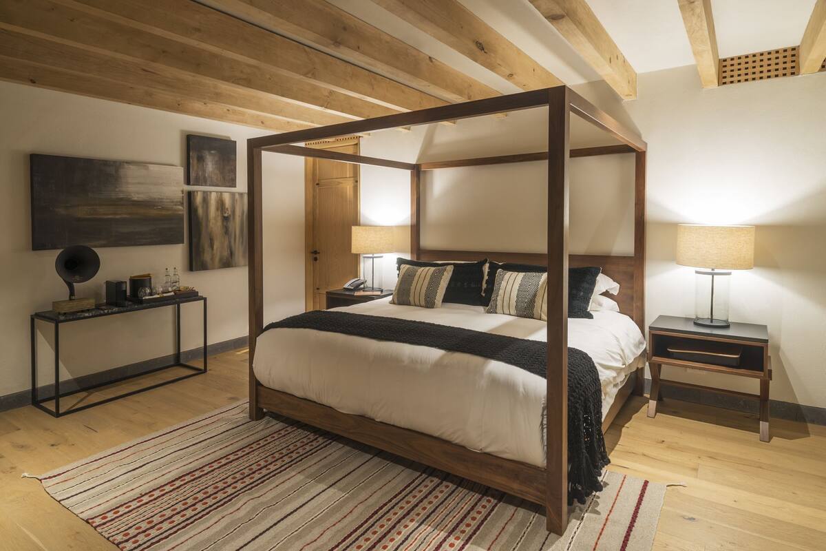 Ein Himmelbett mit Holzrahmen, ein Nachttisch mit Lampe, ein kleiner Tisch ebenfalls mit Lampe und ein Teppich auf dem Boden.