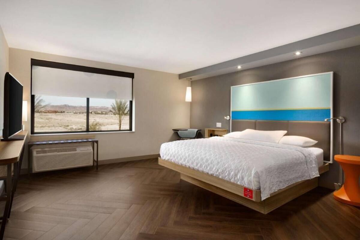 A modern hotel room with a bed, a TV, a desk, and a large window.
