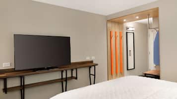 Hotelkamer met een grote flatscreen televisie bevestigd aan een houten console, een bed met een witte dekbedovertrek en een spiegel met een oranje handdoek aan de deur.