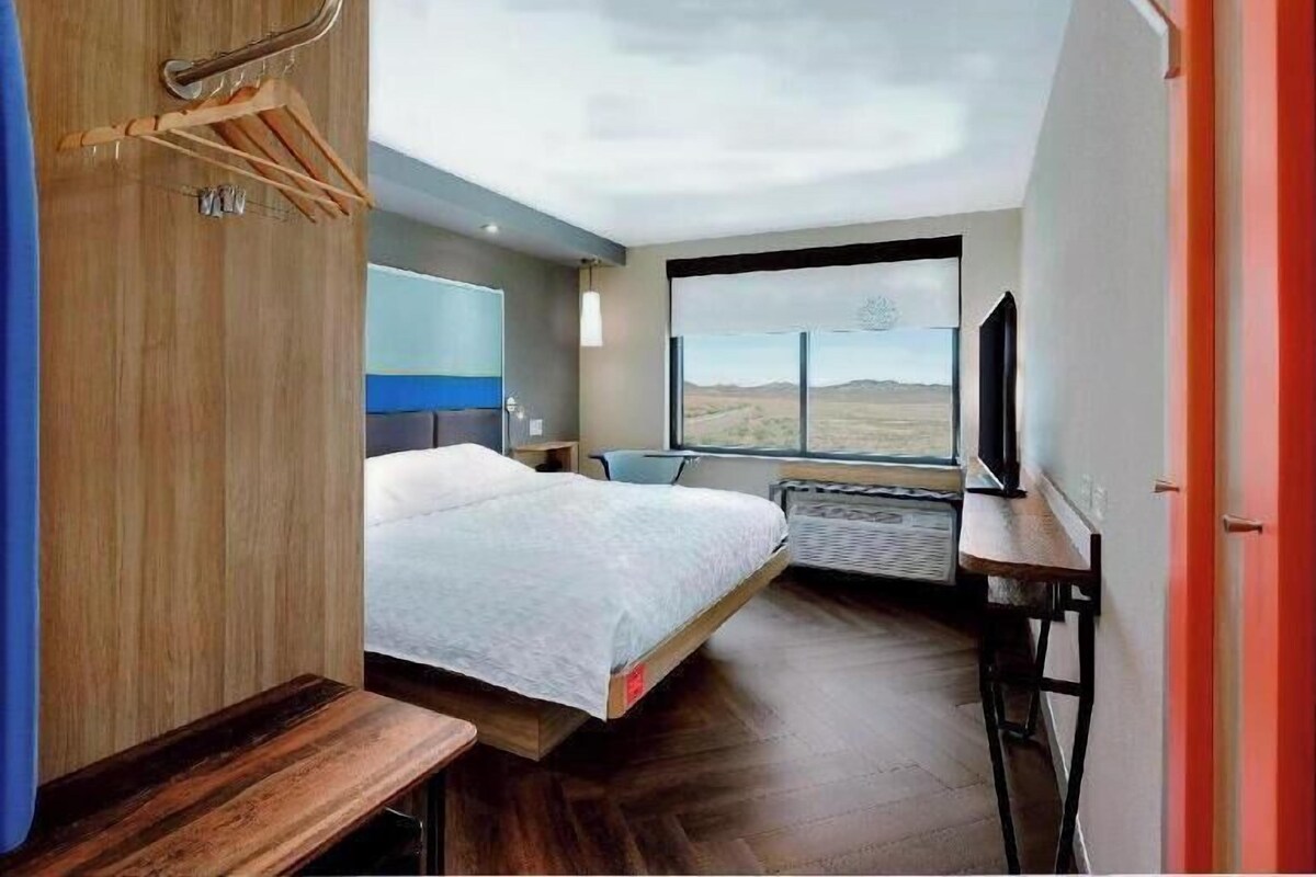 A modern hotel room with a bed, a desk, a TV, and a large window offering a view of the landscape.