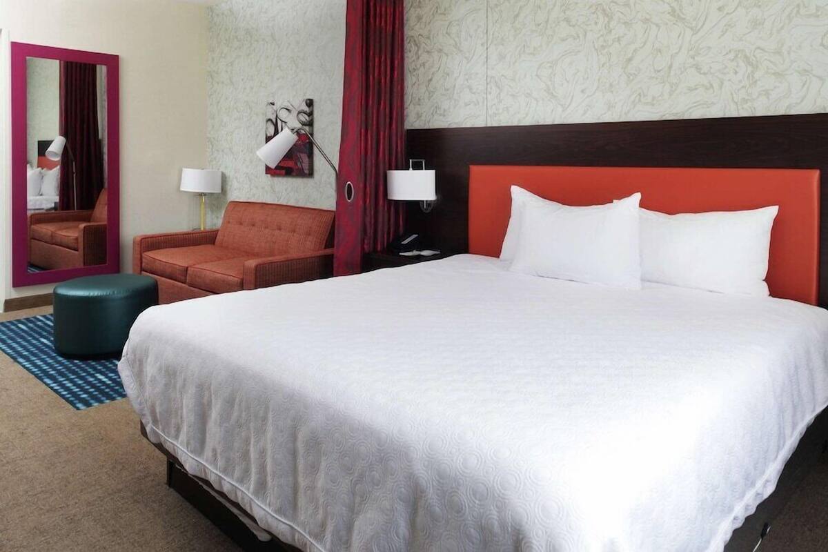 A hotel room with a large bed, a red headboard, two bedside lamps, a mirror, a red curtain, and a patterned wall.