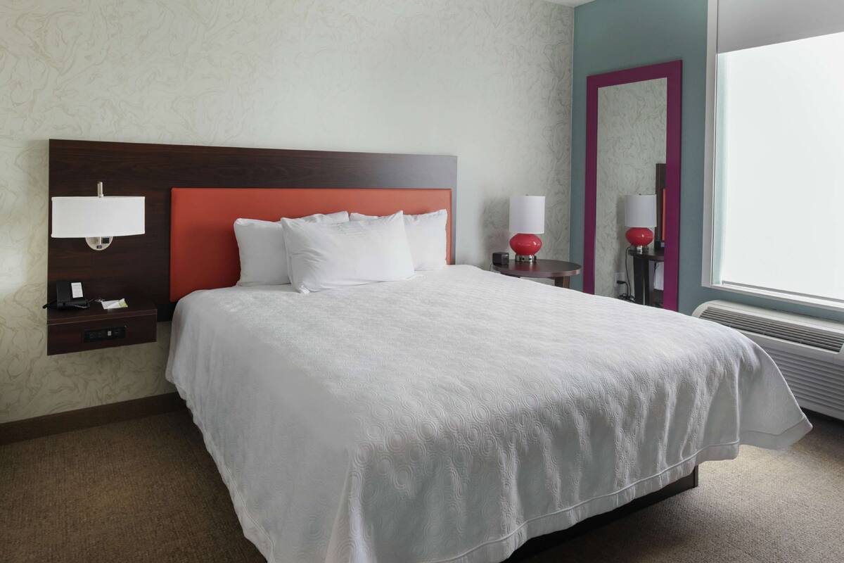 A hotel room with a large bed, a nightstand with a lamp, a mirror, and a window with a view.