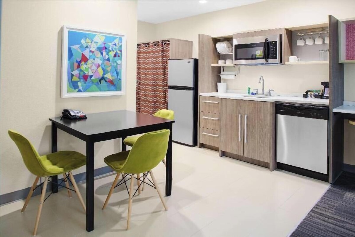 Cocina compacta equipada con refrigerador, microondas, lavaplatos y fregadero. Hay una mesa de comedor con dos sillas.
