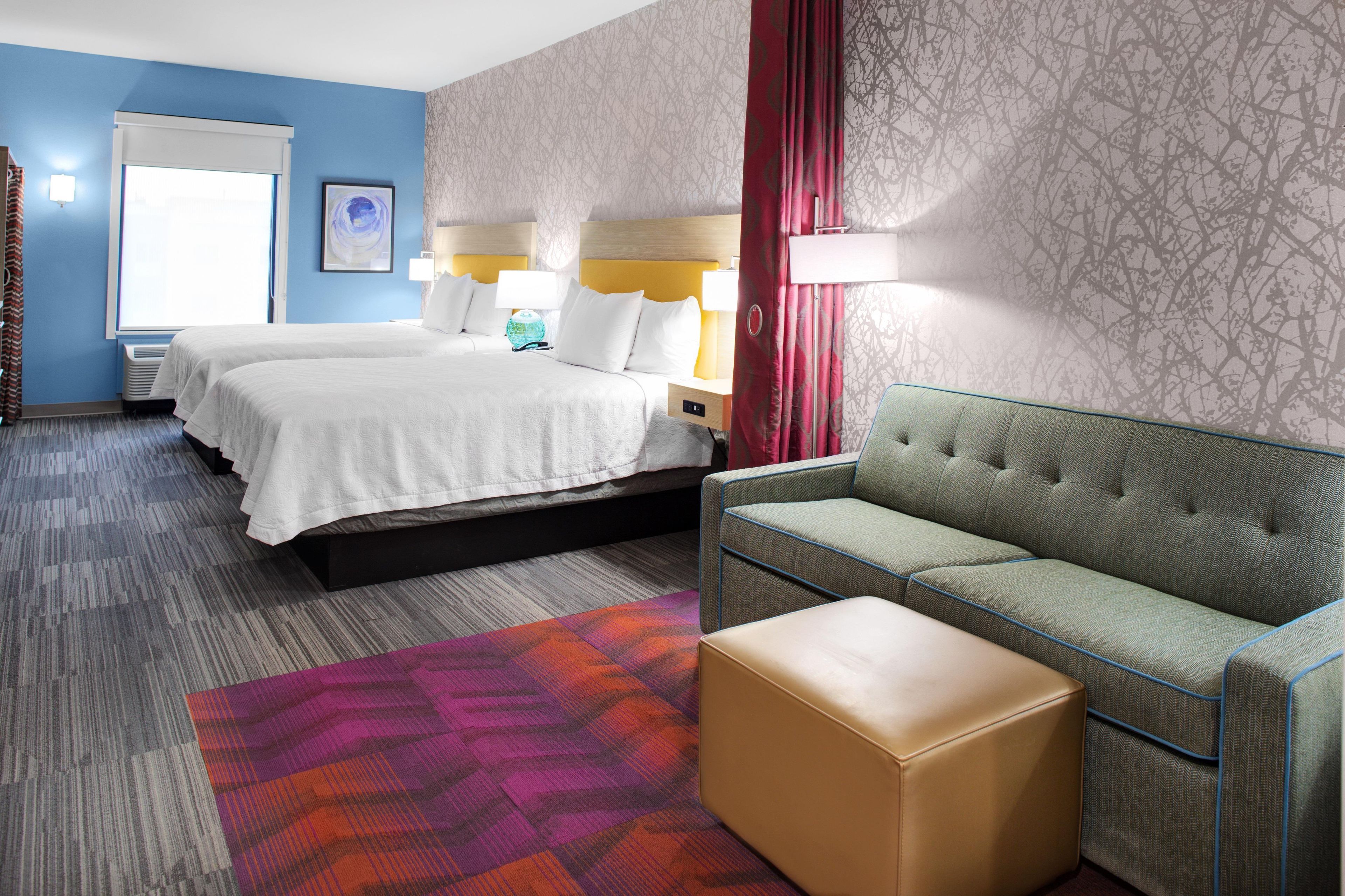 Ein Hotelzimmer mit einem Doppelbett, einem grünen Sofa, einem kleinen Hocker, einem farbenfrohen Teppich und einem Fenster mit Vorhängen.
