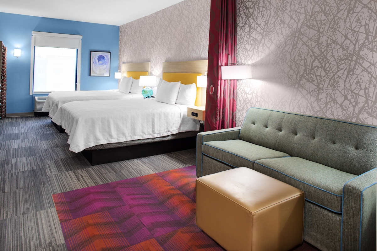 Une chambre d’hôtel avec un lit double, un canapé vert, un petit pouf, un tapis coloré et une fenêtre avec des rideaux.