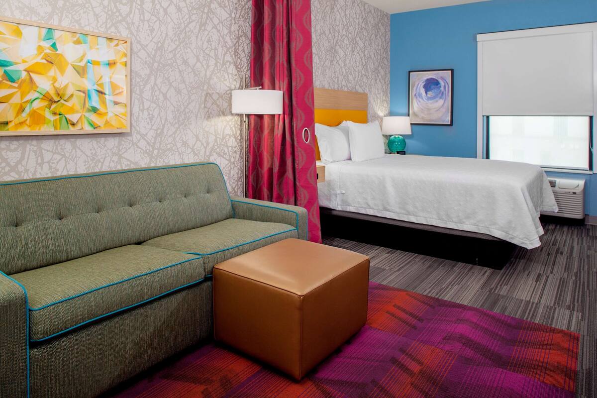 Hotelkamer met een bed, een slaapbank, een klein voetenbankje, een wandlamp, een raam met zonwering en een schilderij aan de muur.