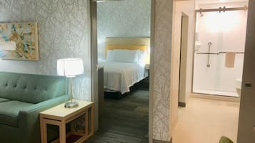 A hotel room with a green sofa, a bedside table with a lamp, a bed with white linens, and a shower area.