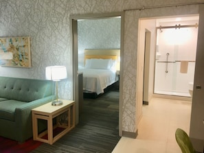 Une chambre d’hôtel avec un canapé vert, une table de chevet avec une lampe, un lit avec du linge de maison blanc et un espace douche.
